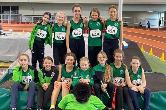 Gruppenzusammenstellung von zw&ouml;lf jungen Athletinnen in Sportkleidung mit Nummern auf einer Leichtathletikanlage.