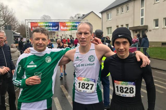 Drei M&auml;nner in Laufbekleidung stehen l&auml;chelnd vor einer Laufveranstaltung, im Hintergrund Zuschauer und ein Zielbanner.