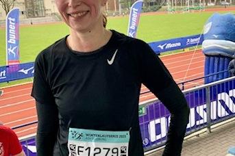 L&auml;chelnde L&auml;uferin in Sportbekleidung mit Startnummer F1279, vor einem Laufstadion im Freien.