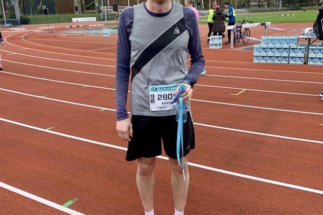 Mann in Sportkleidung mit Medaille steht auf einer Laufbahn, im Hintergrund sind andere L&auml;ufer und Geb&auml;ude sichtbar.