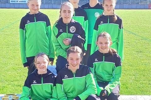 Gruppe von acht Jugendlichen in gr&uuml;nen Sportjacken, l&auml;chelnd auf einem Rasenplatz, umgeben von B&auml;umen.