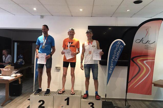Drei Medaillengewinner stehen auf einem Siegertreppchen, jeder h&auml;lt eine Urkunde in der Hand.