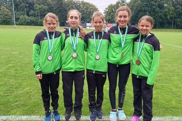 F&uuml;nf M&auml;dchen in gr&uuml;nen Sportjacken mit Medaillen stehen auf einem Fu&szlig;ballfeld und l&auml;cheln in die Kamera.