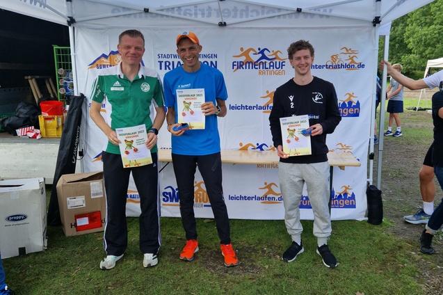 Drei M&auml;nner stehen unter einem Zelt, halten Urkunden in den H&auml;nden und tragen Sportswear nach einem Wettkampf.