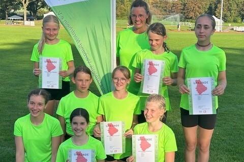 Gruppe von acht M&auml;dchen in neonfarbenen T-Shirts, die stolz Urkunden auf einem Sportplatz halten.