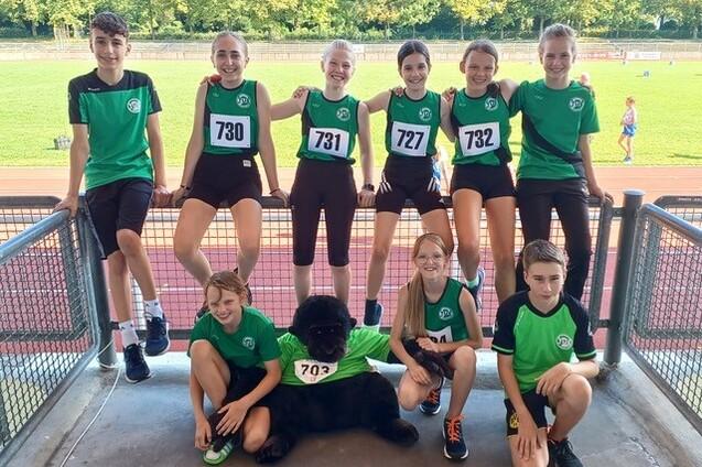 Gruppe von zehn Kindern in Sportkleidung mit Startnummern, posierend mit einem Stofftier auf einem Sportgel&auml;nde.