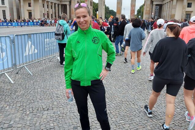 Frau in gr&uuml;ner Laufjacke steht vor dem Brandenburger Tor, umgeben von Teilnehmern eines Lauf-Events.