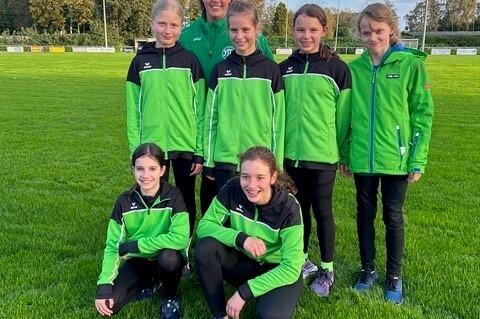 Gruppe von sieben jungen Sportlerinnen in gr&uuml;nen Trainingsjacken auf einer Wiese, l&auml;chelnd posierend.