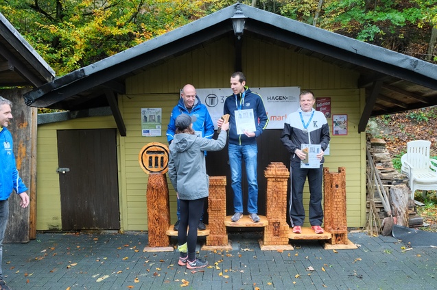 Siegerehrung mit Sportlern auf einem Podest vor einem Holzgeb&auml;ude, jeder h&auml;lt eine Urkunde in der Hand.