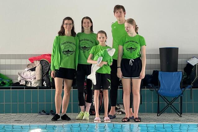 F&uuml;nf Schwimmer in gr&uuml;nen Shirts stehen l&auml;chelnd am Beckenrand eines Hallenbades.