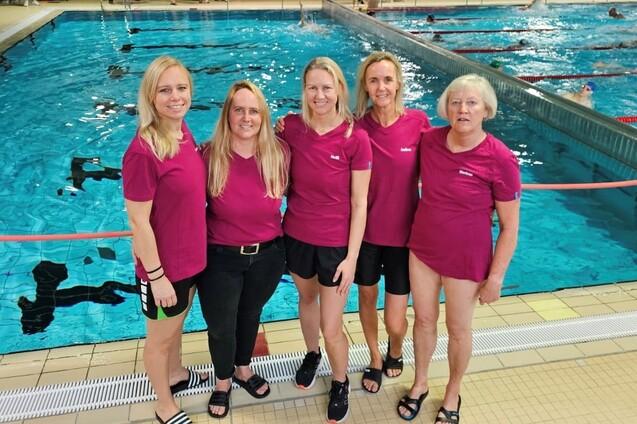 F&uuml;nf Frauen in pinken T-Shirts stehen l&auml;chelnd am Rand eines Schwimmbeckens.