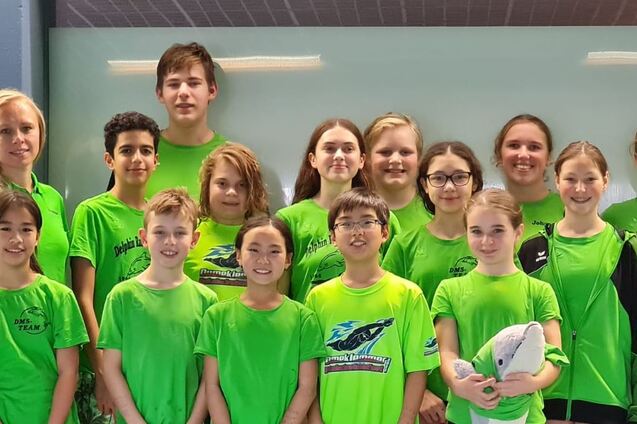 Gruppe von 16 Jugendlichen in gr&uuml;nen T-Shirts, versammelt in einer Sporthalle vor einem hellen Hintergrund.