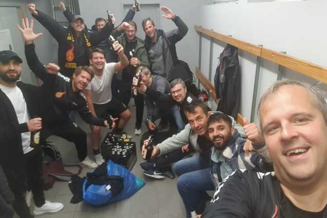 Gruppenfoto von zehn M&auml;nnern in Sportkleidung in einem Umkleideraum, l&auml;chelnd und mit Getr&auml;nken in der Hand.