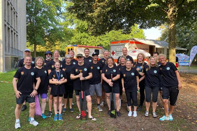 Gruppenfoto von 16 Personen in schwarzen T-Shirts, umgeben von B&auml;umen und einem Rettungswagen im Hintergrund.