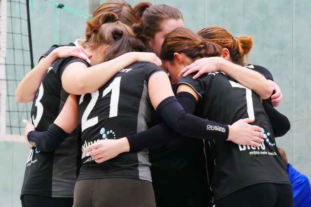 Sechs Volleyballspielerinnen umarmen sich in einer Sporthalle, alle in schwarzen Trikots und gr&uuml;nen Socken.