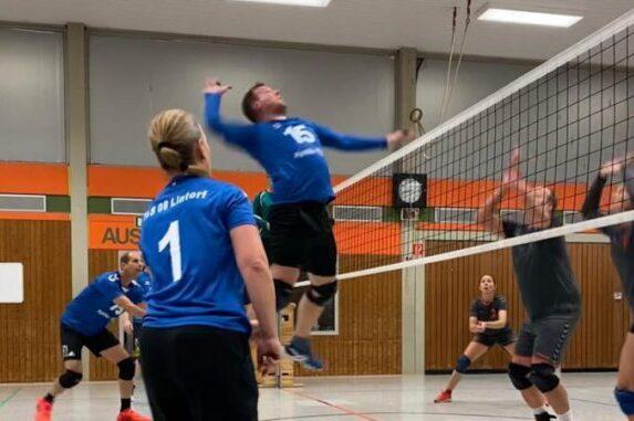 Volleyballspiel: Spieler in blauer Trikotspringt hoch, um am Netz einen Ball zu schlagen, w&auml;hrend andere Spieler attackieren.