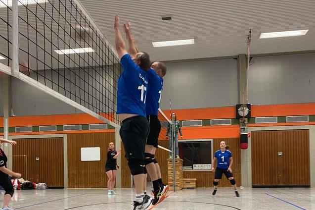 Zwei Volleyballspieler in blauen Trikots springen zum Blocken am Netz, w&auml;hrend andere Spieler im Hintergrund stehen.