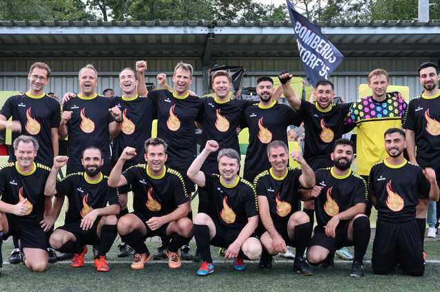 Mannschaftsbild einer Fu&szlig;ballmannschaft in schwarzen Trikots mit Flammenmotiv, alle Spieler posieren l&auml;chelnd auf dem Platz.