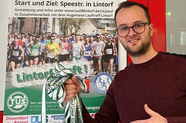 Mann mit Brille zeigt stolz eine Medaille-Anh&auml;ngergruppe vor einem Plakat des Citylaufs Lintorf 2024.
