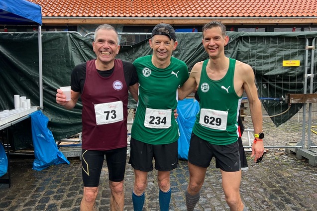 Drei m&auml;nnliche L&auml;ufer in Sportkleidung stehen l&auml;chelnd nebeneinander, vor einem Geb&auml;ude mit rotem Dach.