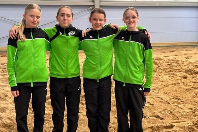 Vier M&auml;dchen in gr&uuml;nen Sportjacken stehen arm in arm vor einer sandigen Fl&auml;che in einer Sporthalle.