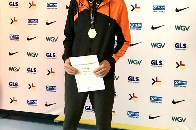 Mann in Sportkleidung mit Medaille und Urkunde vor einem Werbebanner steht stolz l&auml;chelnd.