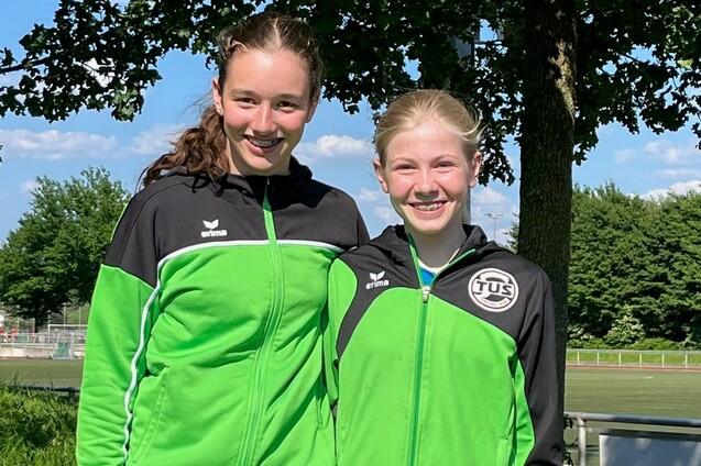 Zwei junge Sportlerinnen in gr&uuml;nen Jacken stehen l&auml;chelnd vor einem Baum auf einer Wiese.