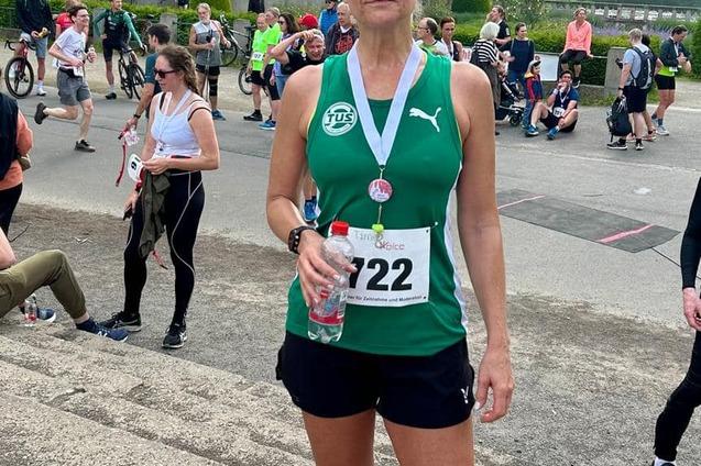 Frau in Sportkleidung mit Medaille und Wasserflasche steht nach einem Lauf im Park, umgeben von anderen Teilnehmern.