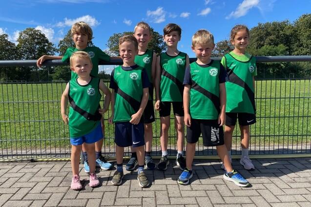 Gruppe von sieben Kindern in gr&uuml;nen Sporttrikots stehen l&auml;chelnd vor einem Sportplatz mit Gras im Hintergrund.