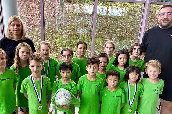 Gruppe von Kindern in gr&uuml;nen Shirts mit Medaillen und einem Ball, zusammen mit zwei Betreuern in einem Raum.