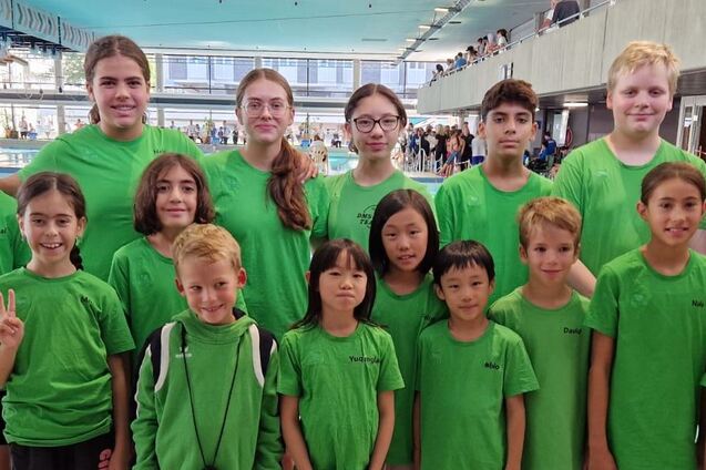 Gruppe von Schwimmern in gr&uuml;nen T-Shirts vor einem Schwimmbecken, l&auml;chelnd und posierend f&uuml;r ein Gruppenfoto.