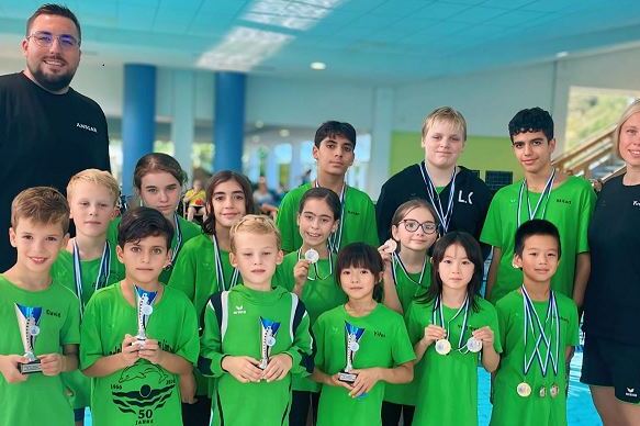 Gruppe junger Schwimmer in gr&uuml;nen Shirts mit Medaillen und Pokalen, posierend nach einem Wettkampf am Schwimmbad.