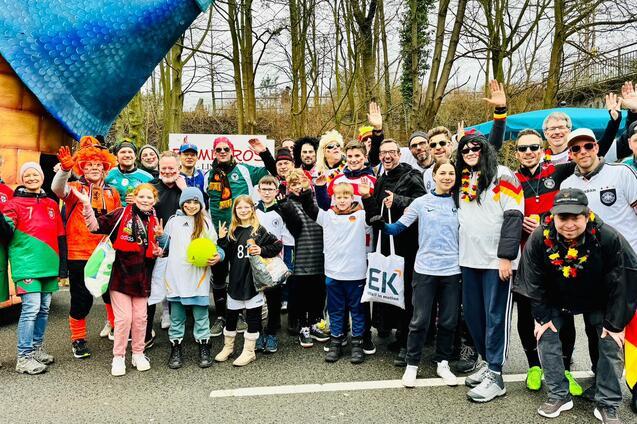Eine Gruppe von Menschen in Farben und Trikots ihrer Nationalmannschaften posiert fr&ouml;hlich auf einer Stra&szlig;e.