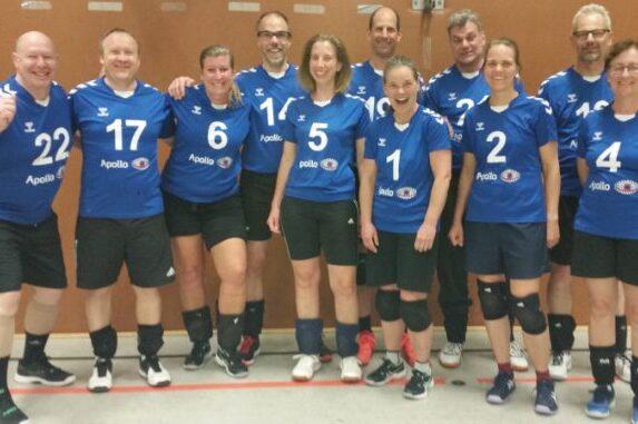 Gruppenfoto einer Volleyballmannschaft in blauen Trikots, l&auml;chelnd, auf einem Hallenboden mit Linien.