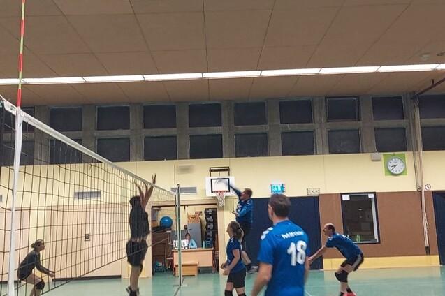 Spielszene in einer Turnhalle mit Volleyballspielern, einer springt zum Blocken am Netz.