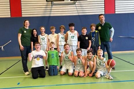 Gruppe von Jungen in Basketballtrikots steht l&auml;chelnd auf einem Sportplatz, umgeben von Trainern. Basketball in der Mitte.