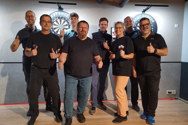 Gruppe von acht Personen steht vor zwei Dartboards in einem Dartsalon und zeigt Daumen hoch.