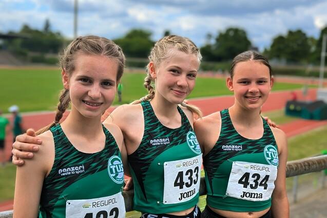 Drei junge Athletinnen in Sportkleidung posieren mit Startnummern auf einer Leichtathletikanlage bei strahlendem Himmel.