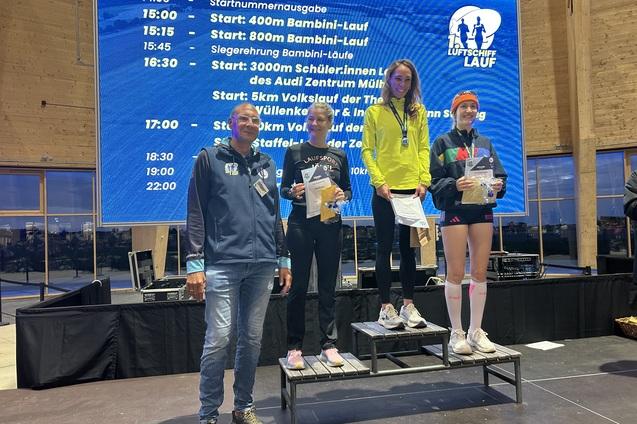 Drei L&auml;uferinnen stehen auf dem Podium mit Medaillen, w&auml;hrend ein Mann neben ihnen applaudiert, Hintergrund mit Veranstaltungstafel.