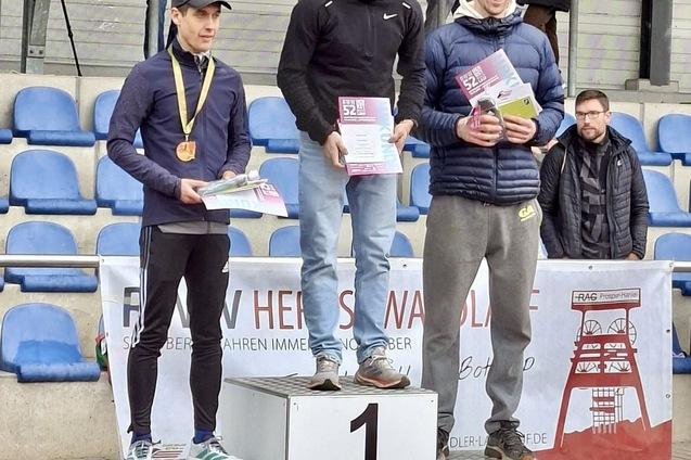 Drei L&auml;ufer stehen auf dem Siegerpodest, halten Urkunden und Medaillen, mit Zuschauern im Hintergrund.