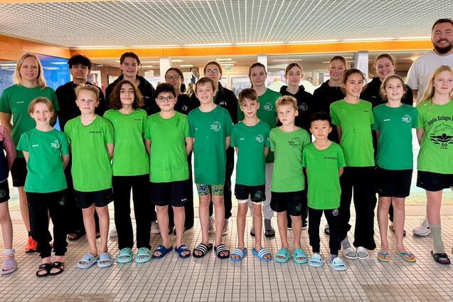 Gruppenshot von Schwimmern in gr&uuml;nen Shirts vor einem Schwimmbecken, begleitet von Trainern und Betreuern.