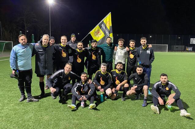 Eine Gruppe von 15 Fu&szlig;ballspielern in schwarzen Trikots posiert mit einer Fahne auf einem Kunstrasenplatz bei Nacht.