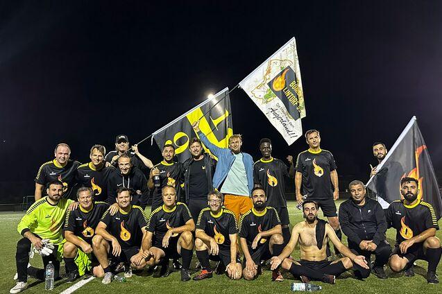 Mannschaft von 20 Fu&szlig;ballspielern in schwarzer Kleidung posiert mit Fahnen auf einem Kunstrasenplatz bei Nacht.