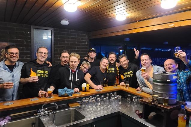 Gruppe von zw&ouml;lf Personen l&auml;chelt an einer Bar, h&auml;lt Gl&auml;ser und posiert vor einem Bierfass.