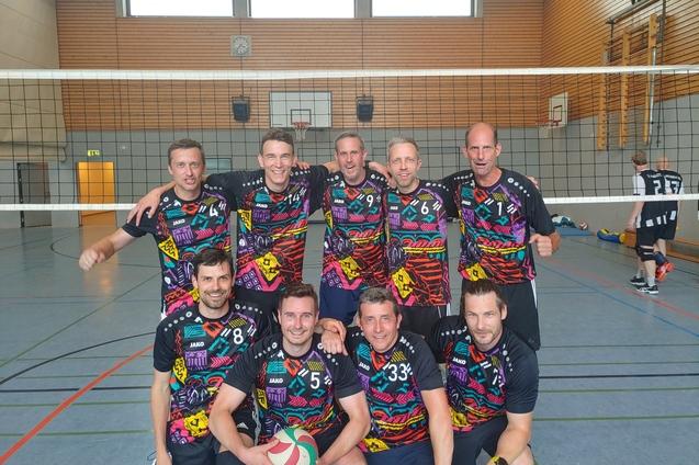 Gruppe von acht m&auml;nnlichen Volleyballspielern in bunten Trikots, posierend vor einem Volleyballnetz in einer Sporthalle.