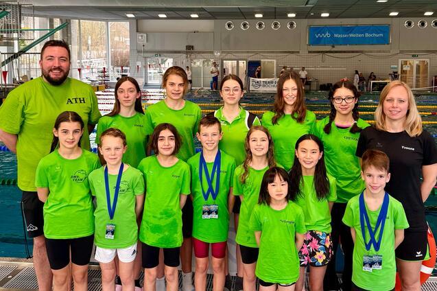 Gruppe von Schwimmern in gr&uuml;nen T-Shirts mit Medaillen, posierend in einem Schwimmbad. Ein Trainer steht hinten.