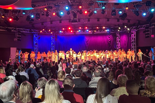 B&uuml;hne mit vielen Personen in orangefarbener Kleidung, bunten Lichtern und Publikum in einem festlichen Veranstaltungsraum.