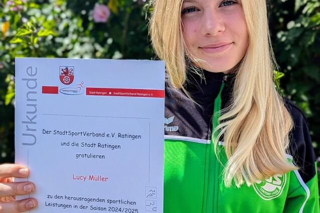 M&auml;dchen in gr&uuml;ner Sportjacke h&auml;lt Urkunde mit dem Text &bdquo;Lucy M&uuml;ller&ldquo; vor einer bl&uuml;henden Hintergrundpflanze.