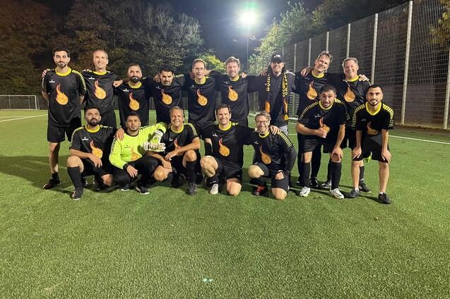 Gruppe von 14 Fu&szlig;ballspielern in schwarzen Trikots mit orangefarbenem Logo posieren gemeinsam auf einem Kunstrasenplatz.