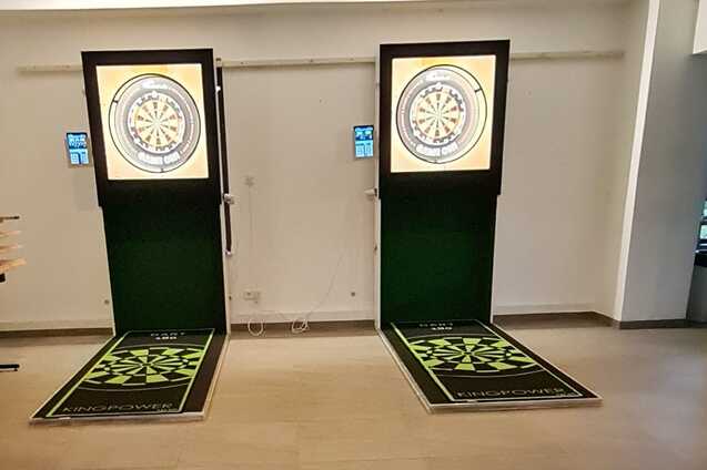 Zwei elektronische Dartscheiben mit farbigen Anzeigen in einem modernen Raum, davor gr&uuml;ne Wurfbereiche.